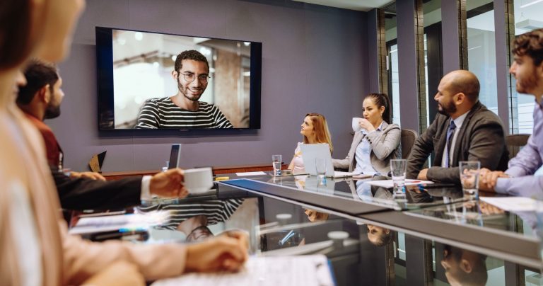 A team in a modern office setting attentively watching and interacting with a colleague on a large screen, indicating a virtual meeting in progress and highlighting the integration of remote participation with in-office work