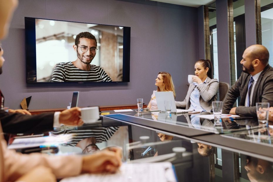 A team in a modern office setting attentively watching and interacting with a colleague on a large screen, indicating a virtual meeting in progress and highlighting the integration of remote participation with in-office work
